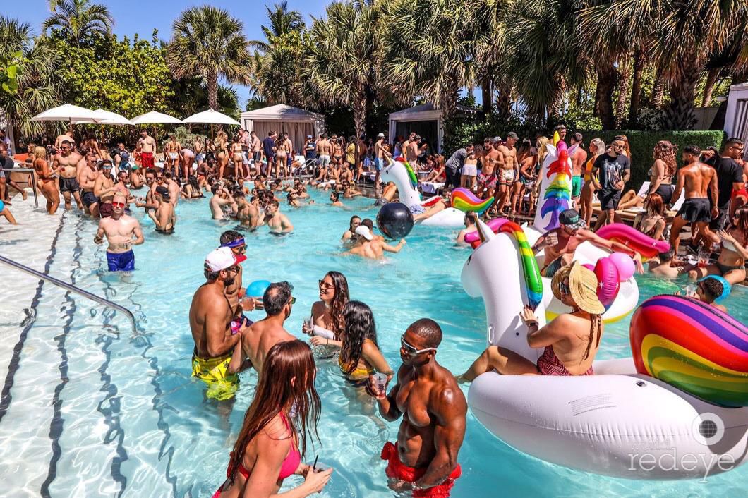 Crowded daytime pool party at a Miami hotel with guests in the water, rainbow pool floats, white cabanas, and palm trees